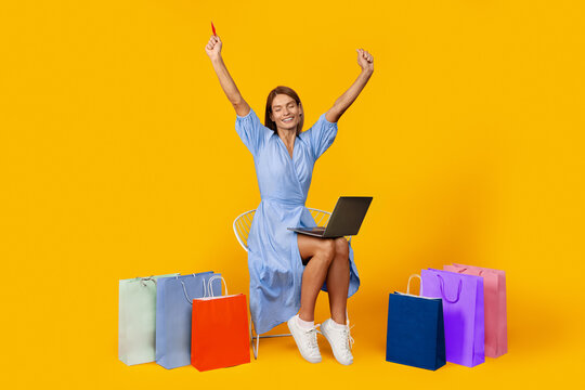 Smiling Young Woman Celebrating Successful Online Shopping With Laptop, Surrounded By Colorful Shopping Bags At Home. Excitement And Joyful Emotion Reflecting Shopping Concept.