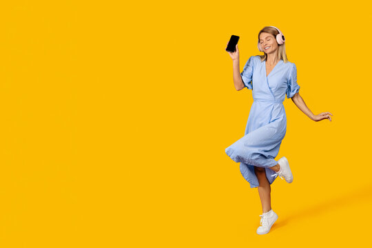 Smiling Young Caucasian Female Enjoying Music While Dancing With Smartphone And Headphones In Studio With Yellow Background, Exuding Positive Energy And Joyful Emotion. Lifestyle Concept.