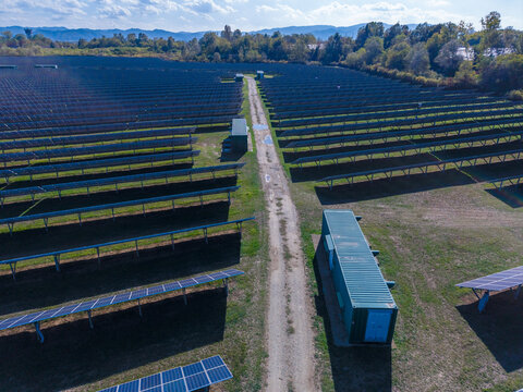 Aerial view of a large solar farm with rows of photovoltaic panels, a central dirt access path, and green storage containers in Torrazza Piemonte, Piemonte, Italy.