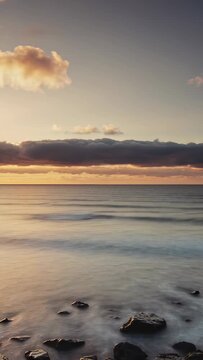 Vertical video: Drifting cloud riding wind heading to low cloud bank above calm sea, showing rocks