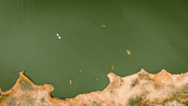 Aerial view of a small white boat with a person floating on the calm green water of a lake near the sandy shoreline in India.
