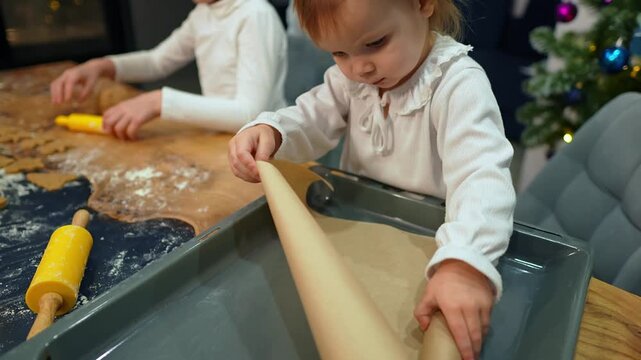 Little girl is unrolling baking paper to put it on sheet for making cookies and her mother is helping. Slow motion of cheerful Caucasian family preparing Christmas pastries together in cozy kitchen