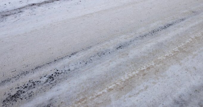 Dirty sand and frozen tyre tracks on winter road surface, horizontal view of slippery slush and road salt on snow covered driving asphalt lane