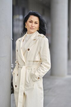 An urban portrait of a woman in a cream trench coat . The scene showcases autumn weather with a city backdrop