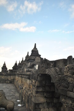 Perspective view along the ancient stone carved walls and walkways of the historic Borobudur Buddhist temple in Java, Indonesia. Features rows of traditional stone stupas against a bright blue sky.   