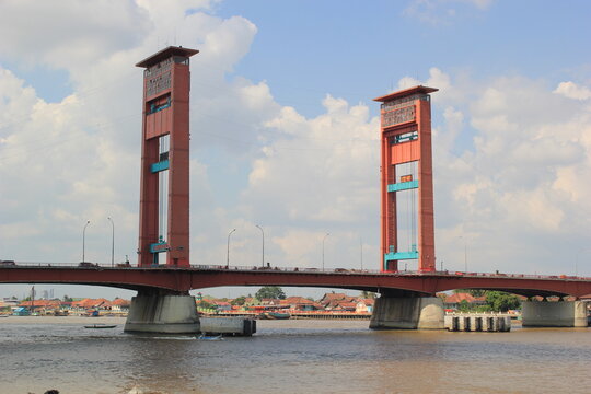 A striking view of the Ampera Bridge, a red vertical lift bridge spanning the wide Musi River in Palembang, Indonesia. The structure features two tall towers with rectangular openings and blue mechani