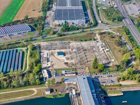 Aerial view of an electrical substation adjacent to a large warehouse, a canal, and solar panel arrays in Hombourg, Grand Est, France.