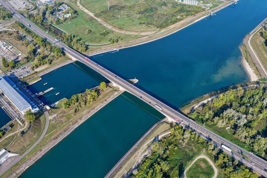 Aerial view of the bridge crossing the Grand Canal d'Alsace, with surrounding lush greenery, calm blue water, and a nearby industrial facility in Hombourg, Grand Est, France.