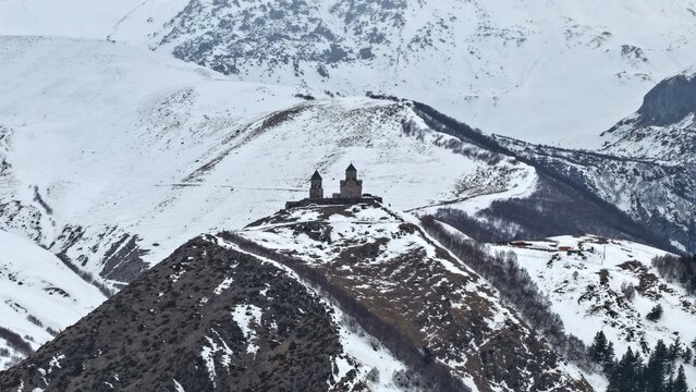 Aerial view of Gergeti Trinity Church perched on a snowy mountain ridge in the Caucasus Mountains, surrounded by snow-covered slopes in Stepantsminda, Mtskheta-Mtianeti, Georgia.