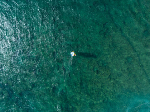 Aerial view of a person wing foiling on the turquoise ocean surface with clear water and visible reef patterns in Maui County, Hawaii, United States.