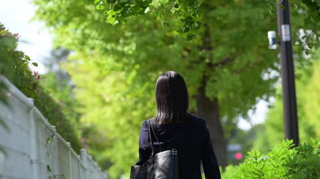 春の新緑に囲まれた屋外を歩く新卒の若い日本人女性社員の後ろ姿、新入社員として新しい社会人生活を始めるビジネネスシーン　顔無しの動画の後姿