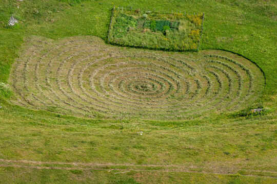 Aerial view of a spiral pattern cut into the green grass of a meadow in the Durmitor mountains, with a small fenced garden nearby in Zabljak, Zabljak Municipality, Montenegro.