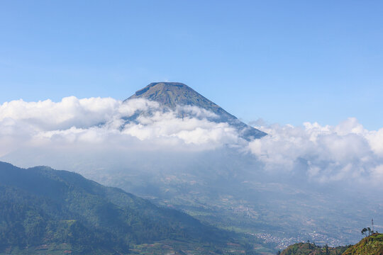 Massive mountain dominates the sky with cloud layers wrapping its peak at Dieng, Kebumen. Vast clouds swirl around rugged slopes, partially veiling the summit. Green hills roll gently below, leading t