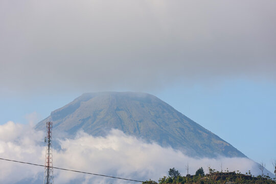 Massive mountain dominates the sky with cloud layers wrapping its peak at Dieng, Kebumen. Vast clouds swirl around rugged slopes, partially veiling the summit. Green hills roll gently below, leading t