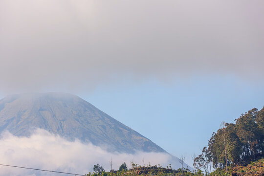 Massive mountain dominates the sky with cloud layers wrapping its peak at Dieng, Kebumen. Vast clouds swirl around rugged slopes, partially veiling the summit. Green hills roll gently below, leading t