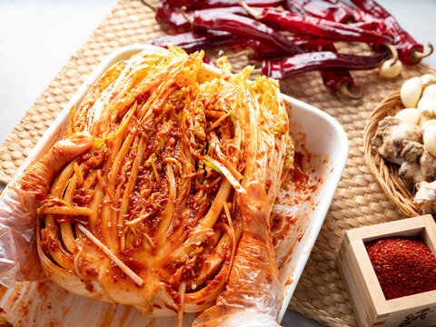 Traditional Korean cabbage Kimchi on a white plate surrounded by raw ingredients like red chili, garlic, ginger, and salt