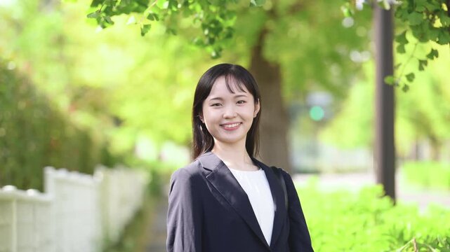 [スロー]春の新緑が美しい屋外をスーツで笑顔で微笑む、新卒の若い日本人女性社員、大学生の就活や新入社員としてフレッシュなイメージのカメラ目線笑顔の動画、髪をかき上げる