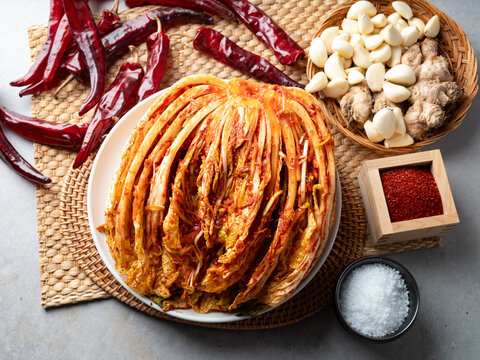 Traditional Korean cabbage Kimchi on a white plate surrounded by raw ingredients like red chili, garlic, ginger, and salt