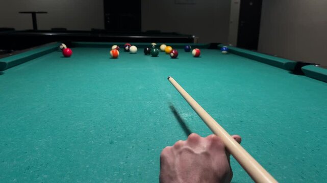 First person view of player striking cue ball with cue stick on billiard table and breaking the rack, sending pool balls scattering in different directions
