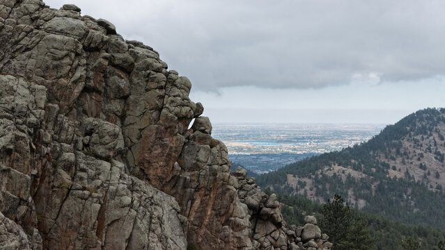 Aerial view of the rocky cliffs of Tram Hill overlooking the expansive valley and distant city under a cloudy sky in Boulder County, Colorado, United States.