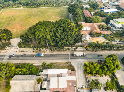 Aerial view of a road with a truck and motorcycles passing by a large green field and lush trees in Klapanunggal, West Java, Indonesia.