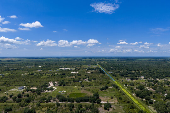 Aerial view of the rural landscape near Lake Okeechobee with a straight canal cutting through green fields, scattered trees, and residential properties, Okeechobee, Florida, United States.