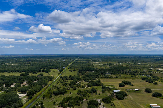 Aerial view of a canal cutting through rural green fields and scattered trees under a bright blue sky with clouds in Okeechobee, Florida, United States.