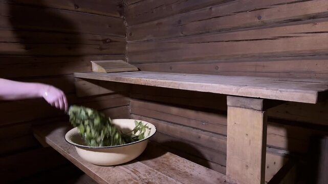 Person shaking a leafy sauna whisk, dipping it into water in an enamel basin, shaking off excess water again and placing the wet whisk on a wooden sauna bench, horizontal video