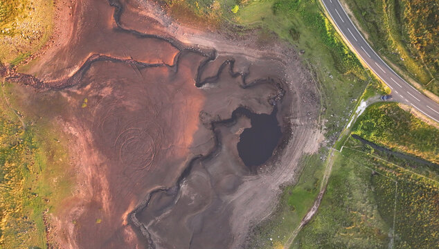 Aerial view of Loughareema, the vanishing lake, showing dark water, textured muddy ground, and a winding road in Ballycastle, Northern Ireland, United Kingdom.