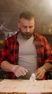 Vertical video. Joinery workshop. Wood gluing. Male carpenter in gloves applying adhesive with brush to set of sanded planks furniture in smoky interior.