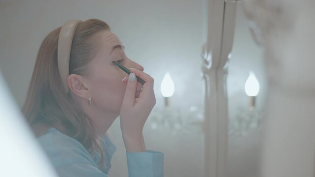 Woman applies mascara while looking into mirror. She wears light blue shirt and headband. Focus is on her careful, precise movements. Scene shows personal grooming routine. Soft lighting adds calm