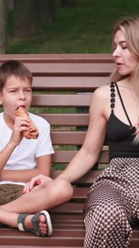 Vertical video. Family snack. Park eating. Relaxed mother smiling son together on brown wooden bench enjoying delicious pastry sharing food outside.