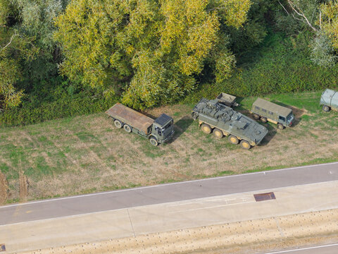 Aerial view of military vehicles including a flatbed truck and an armored personnel carrier parked on a grassy verge next to a road in the United Kingdom.