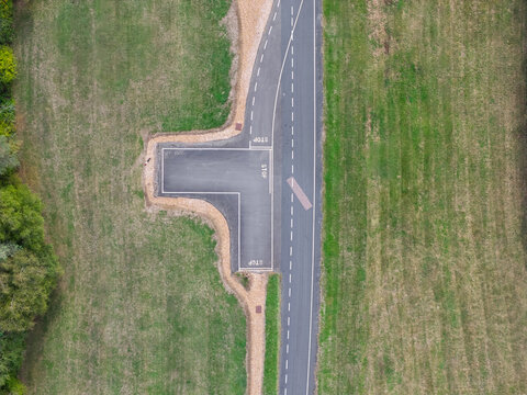 Aerial view of a vehicle testing track section with asphalt road, white stop markings, and surrounding green grass fields in United Kingdom.
