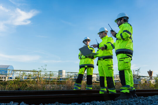 Industrial engineers using laptop and radio for inspection near factory railway tracks, Team of industrial inspectors reviewing data on laptop near chemical plant facility