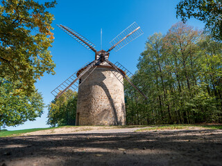 Front view of the historic 19th-century stone windmill in Holic, Slovakia, with four wooden sails. Traditional milling heritage and rural architecture surrounded by trees under a clear blue sky. © Marek