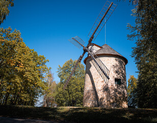 Historic 19th-century stone windmill in Holic, Slovakia, seen from the side among trees under a clear blue sky. Traditional wooden sails, rural architecture and preserved milling heritage. © Marek