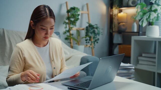 Young Asian woman reviews financial documents, bills and expenses on laptop at home. Personal finance budget planning, Tax invoice paperwork review, Household savings and expense management concept.