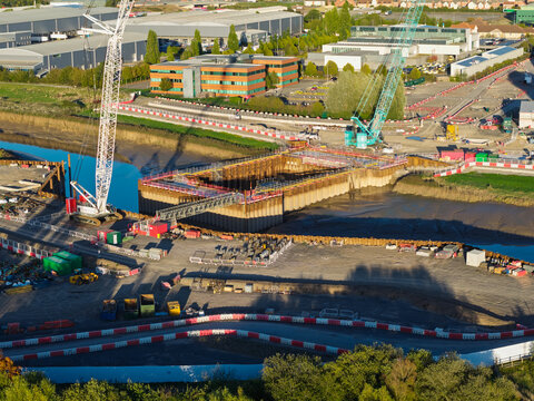 Aerial view of flood defence barrier construction featuring large cranes and metal sheet piling along the riverbank in the United Kingdom.