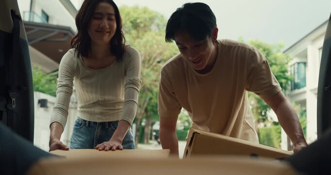 Young Asian couple unloads cardboard box from car to move into house. Family relocation home mortgage loan, Real estate vehicle transport delivery, Moving day logistics, New home arrival pack concept.