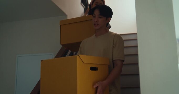 Young Asian couple carries boxes and home decor through bright room while moving house. Relocation teamwork, unpacking furniture, new home setup, interior decorating and shared lifestyle concept.