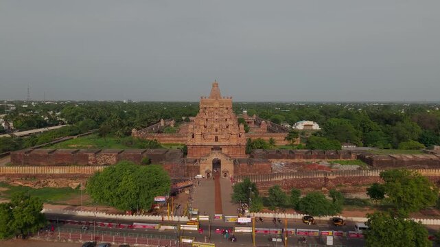 Aerial drone view of Brihadeeswara Temple entrance with monumental stone gateway, ancient Chola architecture and tree lined grounds beside a busy road in Thanjavur, Tamil Nadu, India.