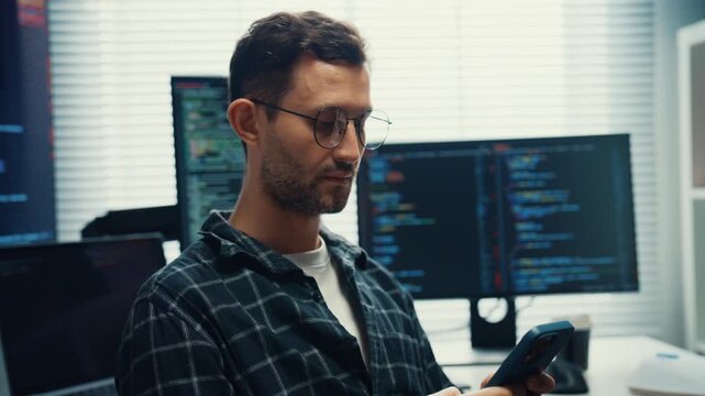 Young Caucasian male developer use mobile phone while working with multiple monitor in office. Professional AI developer workspace, Secure computer network system, Advanced cybersecurity data concept.