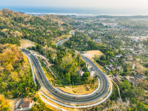 Aerial view of the winding Kelok 23 road featuring sharp curves through lush hills, dense tropical vegetation, and nearby rural houses in Kecamatan Kretek, Daerah Istimewa Yogya, Indonesia.