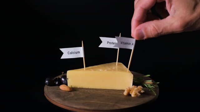 A male hand places protein, calcium, vitamin A and zinc labels into cheese on a wooden stand against a black background. Cheese nutrition concept