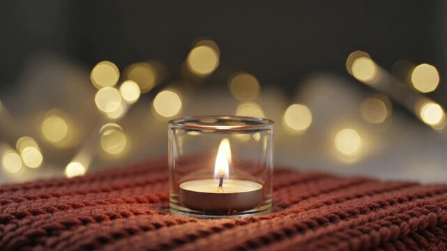 Close-up macro shot of a small tealight candle burning in a glass holder resting on a cozy rusty brown knitted woolen sweater with soft blurred bokeh lights in the background.