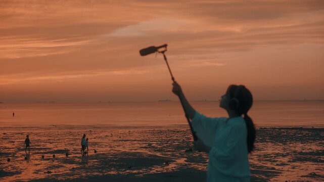 Woman Holding Shotgun Microphone and Headphones Recording Nature Ambient Sound on Mudflat at Sunset