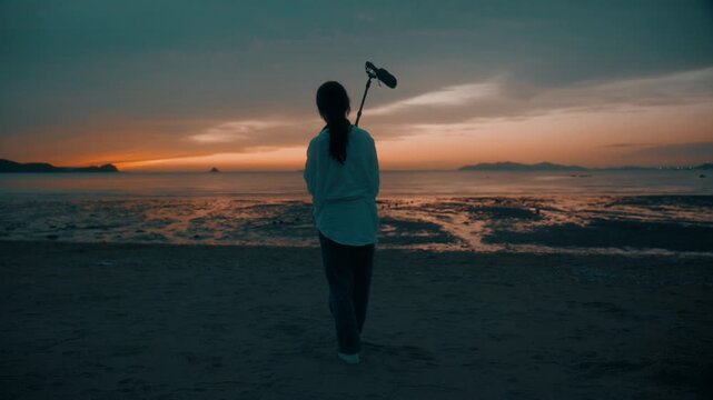Full Shot Rear View of Woman Walking towards Water with Shotgun Microphone on Mudflat at Sunset