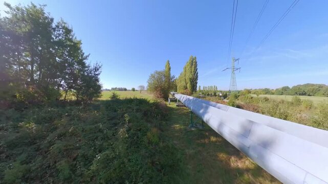 Low angle reverse FPV shot reveals a large industrial pipeline running through rural landscape with electric pylons above, suitable for energy infrastructure, industrial transition and changing energy
