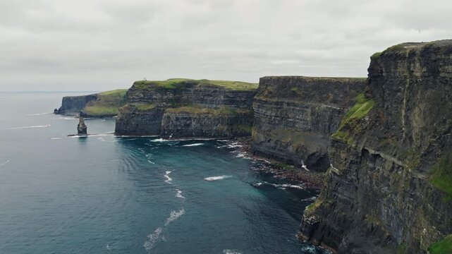 Overcast aerial view of layered sea cliffs at Cliffs of Moher in County Clare, Ireland. A rock stack and white wave foam mark this Atlantic coast sightseeing landscape.